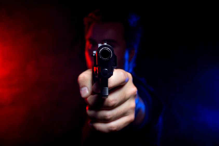 A man with a focused expression holds a handgun towards the viewer. Dramatic lighting creates an intense atmosphere with contrasting red and blue hues.